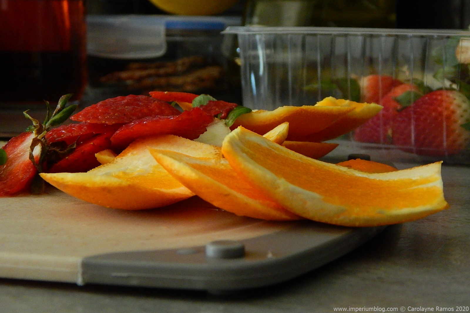 fotografia de cascas de laranja e morangos, sobre uma tábua de cortar de cozinha. ao fundo, uma caixa de plástico com alguns morangos inteiros. imagem alusiva a um artigo acerca da reutilização de cascas de alimentos.